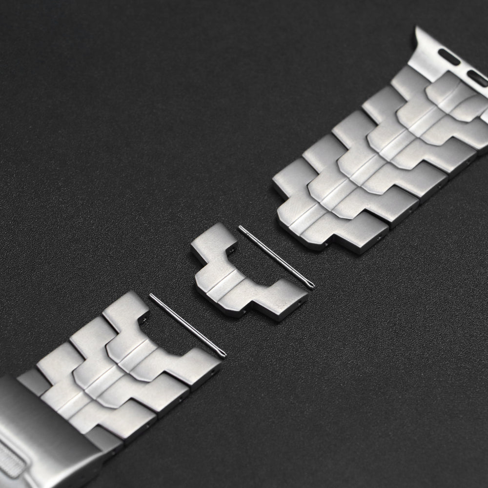 Close-up van een gedemonteerde zilverkleurige metalen horlogeband met geometrische schakels op een donkere achtergrond. Een losse schakel en twee metalen pinnen liggen tussen de twee delen van de band in, wat het proces van het inkorten van het horlogebandje toont.