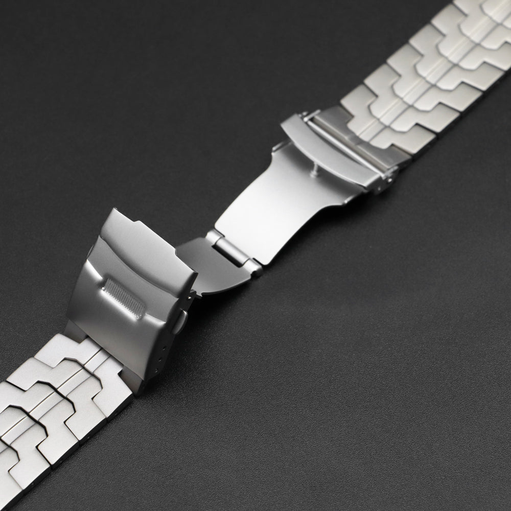 Close-up van een geopende zilverkleurige metalen vouwsluiting van een horlogeband met geometrische schakels, liggend op een donkere achtergrond.