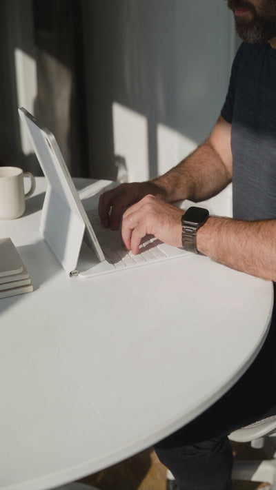 Een man typt op een witte tablet met toetsenbord aan een ronde witte tafel, beschenen door fel zonlicht. Aan zijn pols draagt hij een smartwatch met een donkergrijze of zwarte metalen schakelband. Op de achtergrond op tafel staan een witte koffiemok en enkele notitieboekjes.