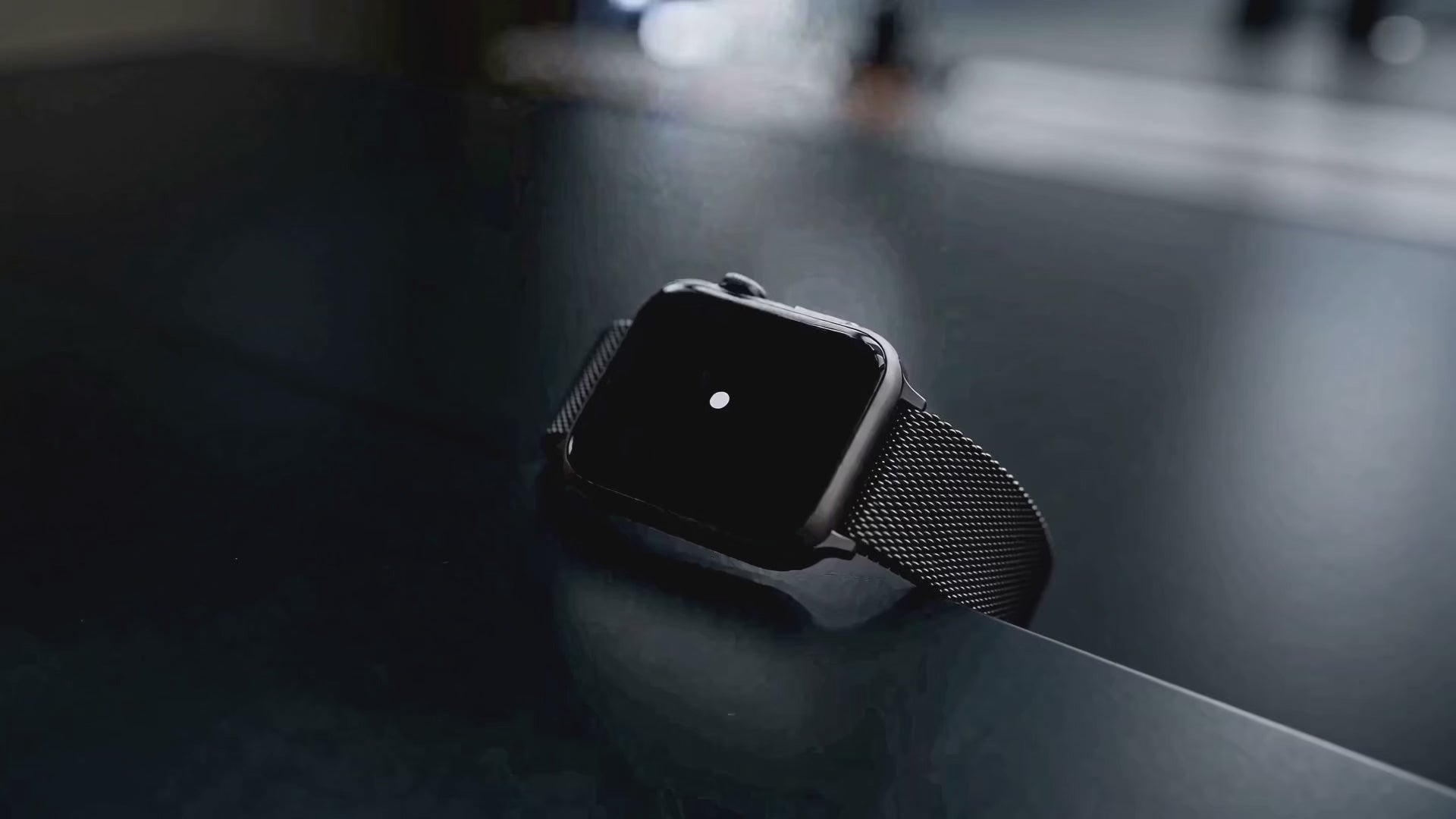 Een close-up van een smartwatch met een zwarte metalen Milanese horlogeband, liggend op een donker en reflecterend oppervlak. Het scherm van het horloge is zwart met een kleine witte stip in het midden, vastgelegd in een sfeervolle, donkere belichting.
