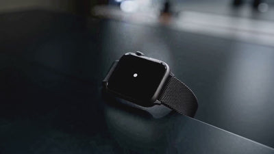Een close-up van een smartwatch met een zwarte metalen Milanese horlogeband, liggend op een donker en reflecterend oppervlak. Het scherm van het horloge is zwart met een kleine witte stip in het midden, vastgelegd in een sfeervolle, donkere belichting.